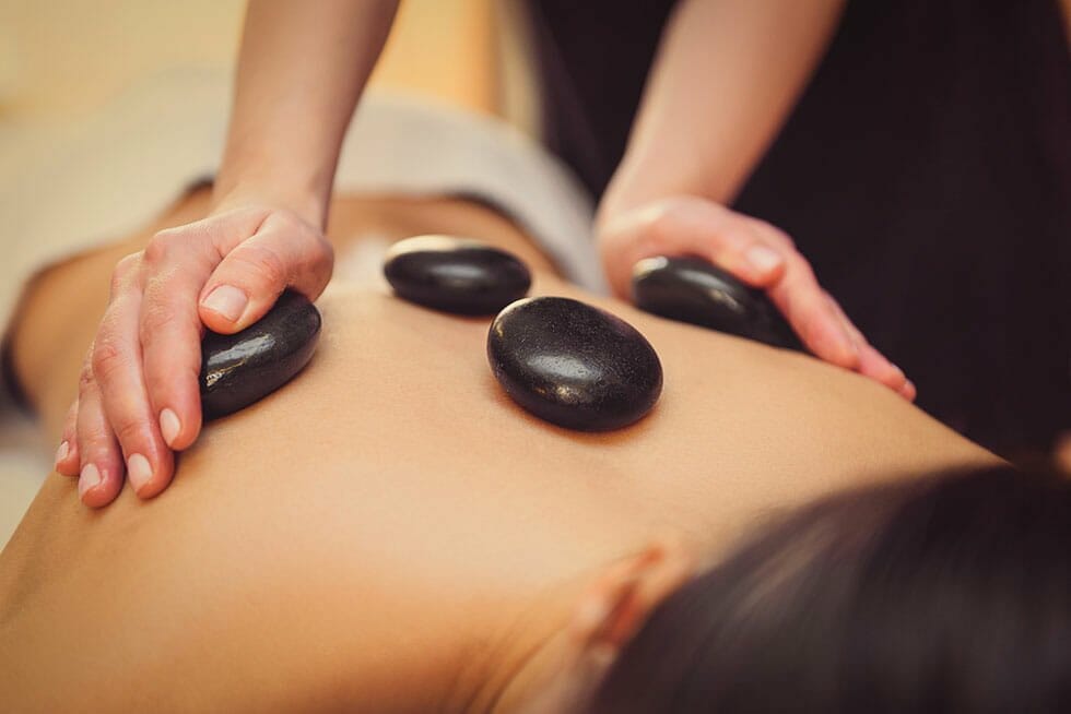 Hot-Stone-Massage im Sonnenhof. Erwärmte, glatte Steine werden fachmännisch auf dem Rücken des Gastes platziert. Dieser genießt die Anwendung entspannt.